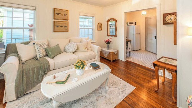 A cozy living room with a white sofa, a coffee table with books and flowers, and a wooden floor. There are framed pictures on the wall and a clock on the wall. - Stay Vintage at Dandelion (McKinney)
