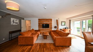 A modern living room with a large sectional sofa, a flat-screen TV mounted on a wooden wall, and a large window providing natural light. - Residence Alpin TOP 1 Incl Summer Card (Kaprun)