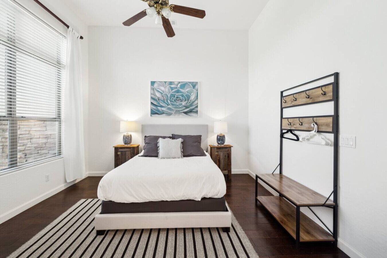 A bedroom with a bed, a ceiling fan, a wall-mounted coat rack, and a window with blinds.