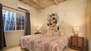 Une chambre à coucher avec un grand lit, deux tables de chevet, un climatiseur fixé au mur et une fenêtre avec des rideaux.