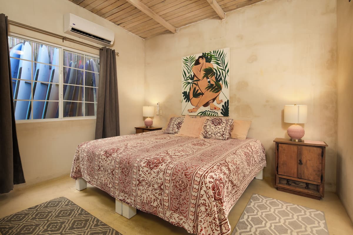 Ein Schlafzimmer mit einem großen Bett, zwei Nachttischen, einer Wand-Klimaanlage und einem Fenster mit Vorhängen.