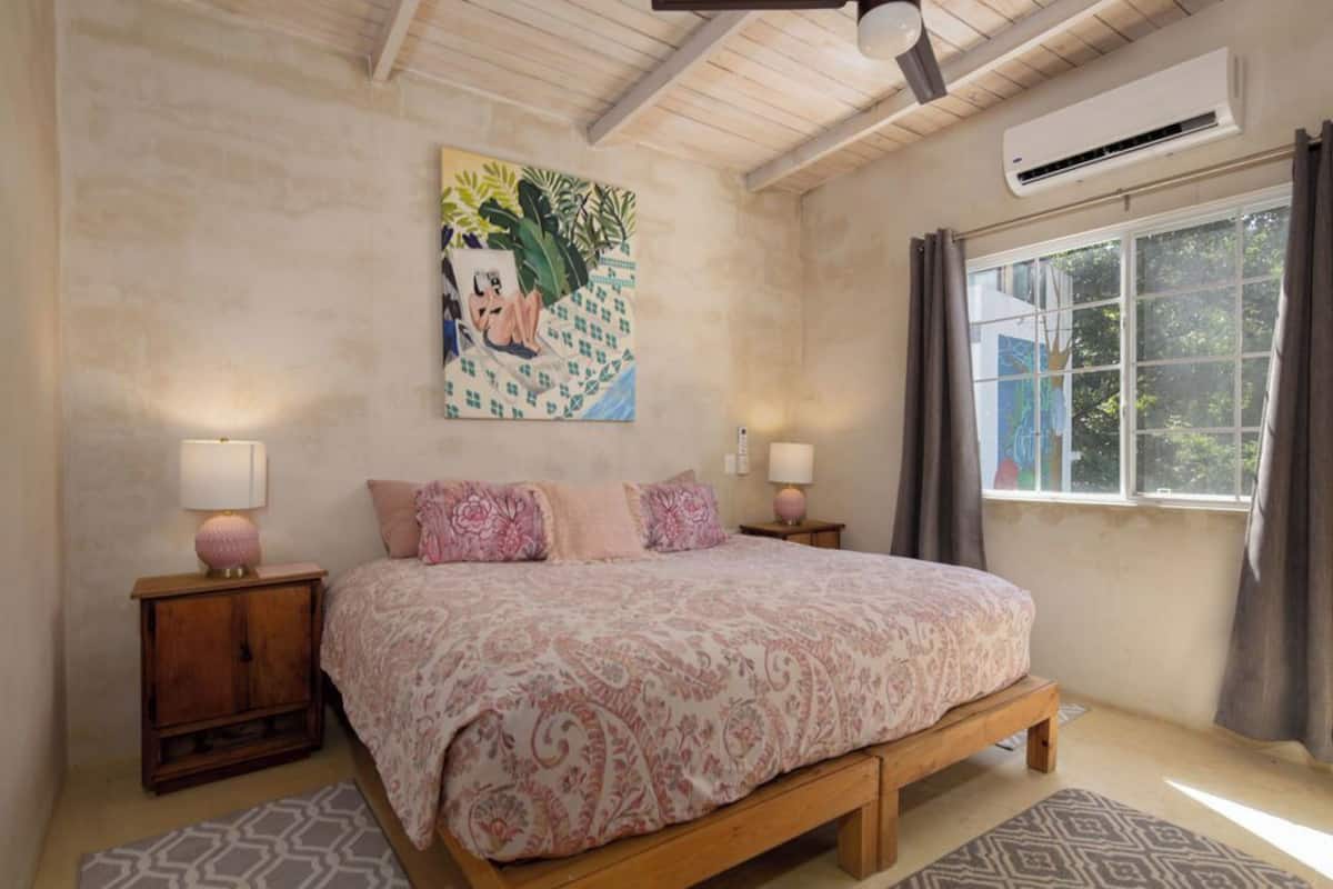 Ein Schlafzimmer mit einem Bett, zwei Nachttischen, einem Deckenventilator, einer Klimaanlage, einem Fenster mit Vorhängen und einem Wandbild.
