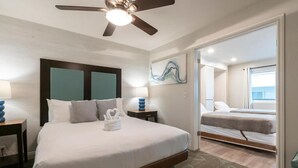 Une chambre d’hôtel avec un grand lit, un ventilateur de plafond, une table de chevet avec une lampe et une vue sur une autre chambre.