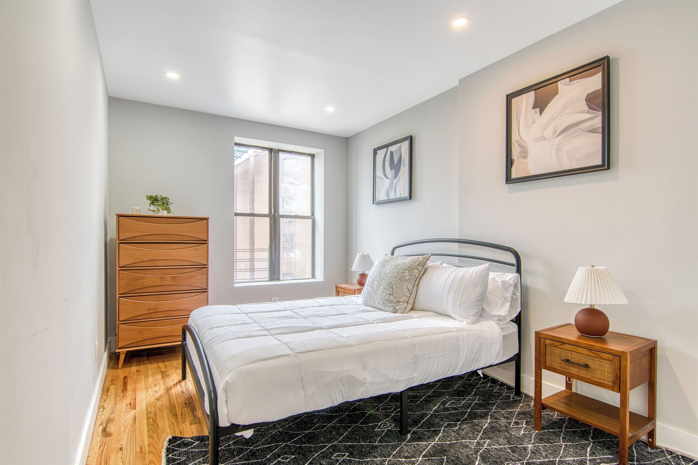 Une chambre à coucher avec un lit, une commode, une table de chevet avec une lampe, une fenêtre et un tableau au mur.