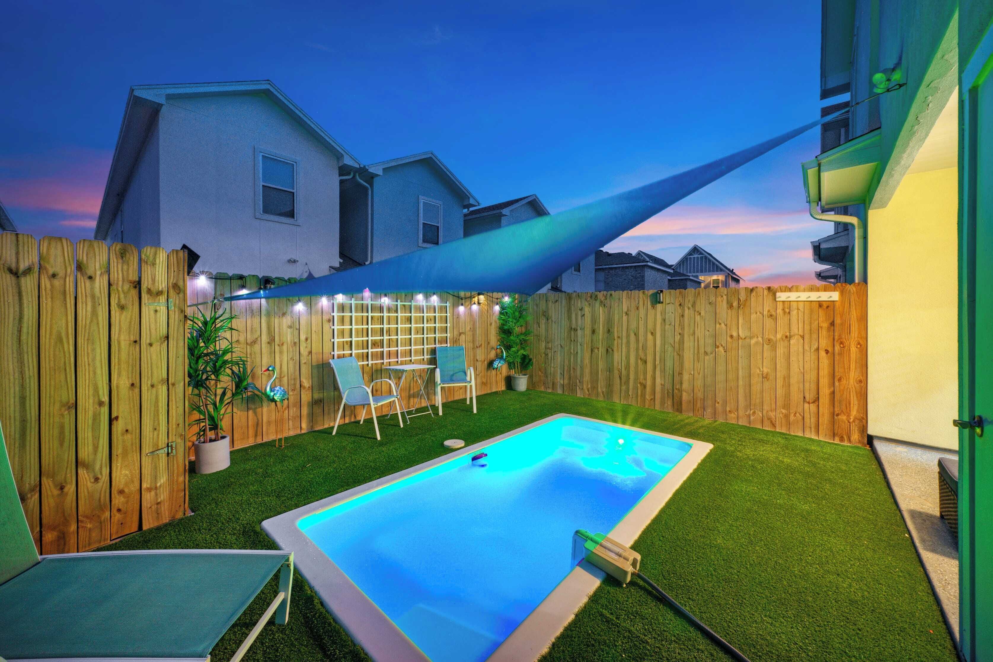 Un jardin avec une piscine, une pelouse artificielle, une clôture en bois et un espace patio aménagé avec des chaises et une table.