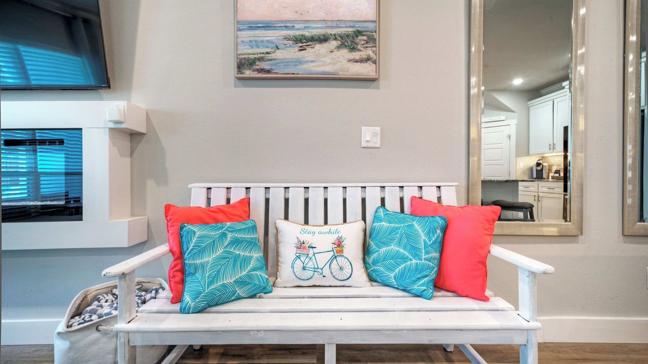 Un banc blanc orné de coussins aux motifs rouges et bleus, un tableau représentant une scène de plage et un miroir reflétant une partie de la cuisine.