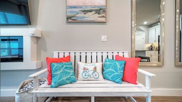 A white bench with red and blue patterned cushions, a painting of a beach scene, and a mirror reflecting a kitchen area.