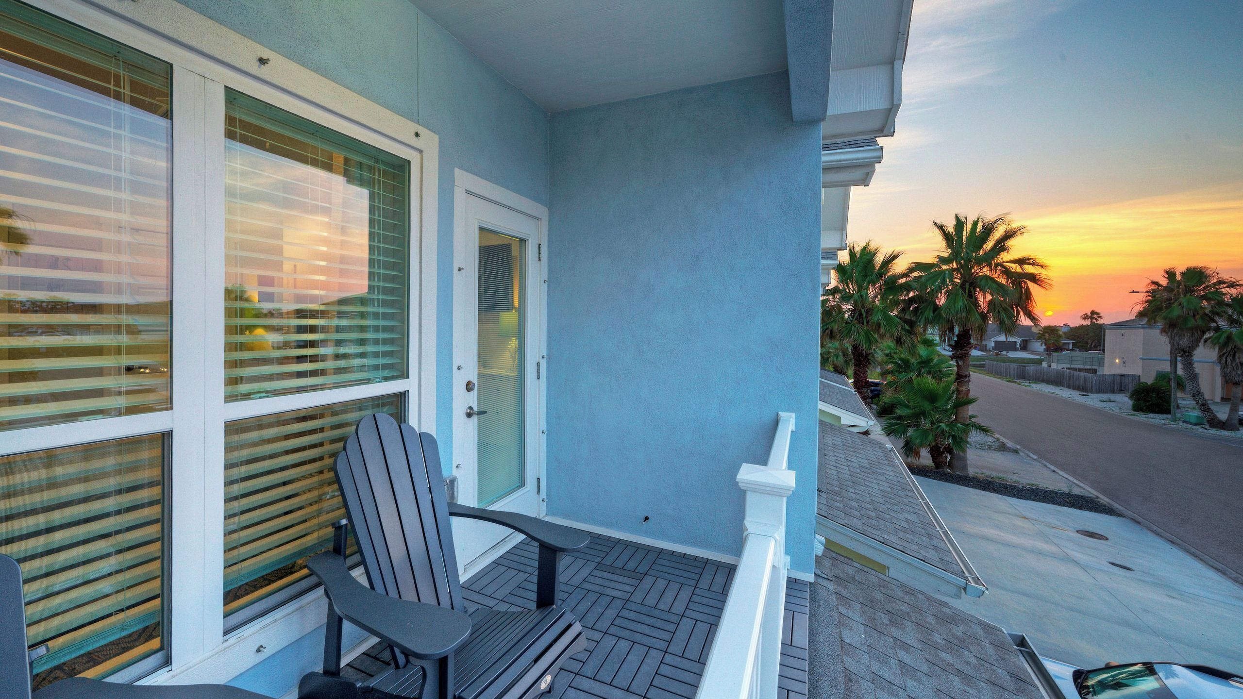 Un balcon avec une chaise, une porte et une vue sur un coucher de soleil au-dessus d’une rue résidentielle.