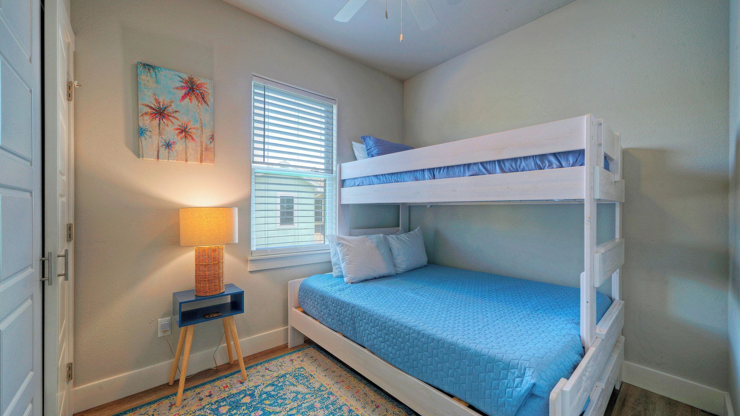 A bedroom with a bunk bed, a bedside lamp, a window with blinds, and a painting on the wall.