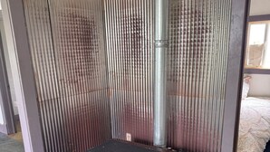 Une pièce avec un poêle à bois, une fenêtre et une grille d’aération en métal.