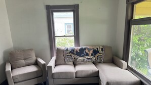 Une pièce d’angle avec un canapé et un fauteuil de couleur beige, un plaid à motifs et une vue par la fenêtre.