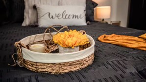 Una cama con un edredón oscuro estampado, una almohada de bienvenida, una cesta tejida con una vela y flores, y una manta al lado.