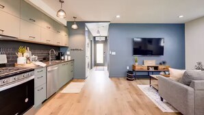 Una cocina moderna con armarios grises, estufa integrada e iluminación colgante. Se observa un área de estar con un sofá gris y un televisor de pantalla plana.