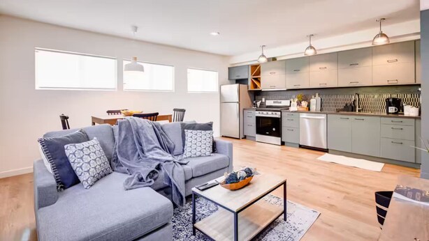 Un apartamento moderno con un sofá gris, una mesa de centro y una cocina con armarios grises y electrodomésticos de acero inoxidable.