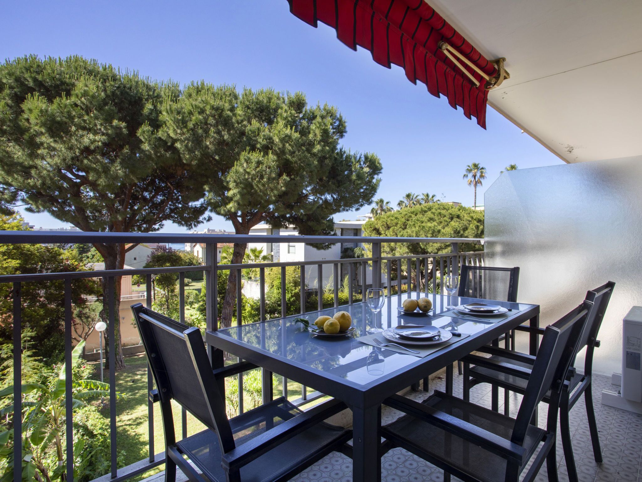 A balcony with a glass table set for dining, surrounded by metal railings, and overlooking a garden with trees and buildings.