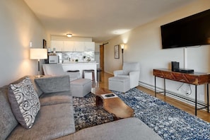 A modern living room with a sofa, chairs, a coffee table, a TV, and a kitchen in the background.