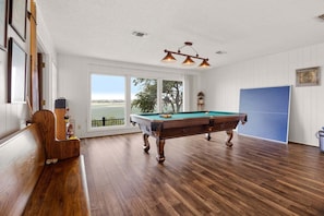 Uma mesa de bilhar em um espaço com piso de madeira, janelas amplas e vista para o exterior.