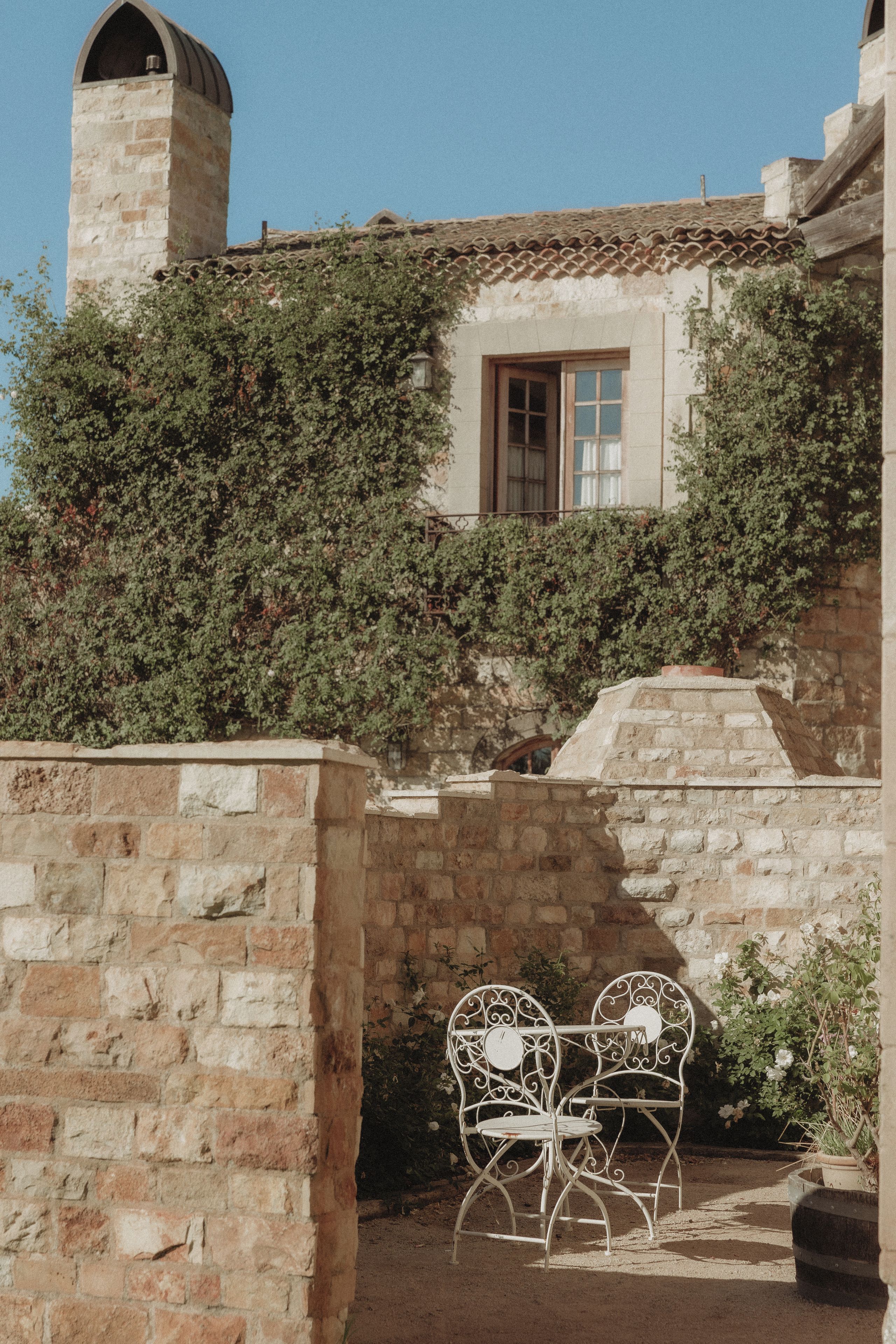 Un muro de piedra con una silla y una mesa de metal, una ventana que da a una vista de un edificio con tejado de azulejos y una chimenea.
