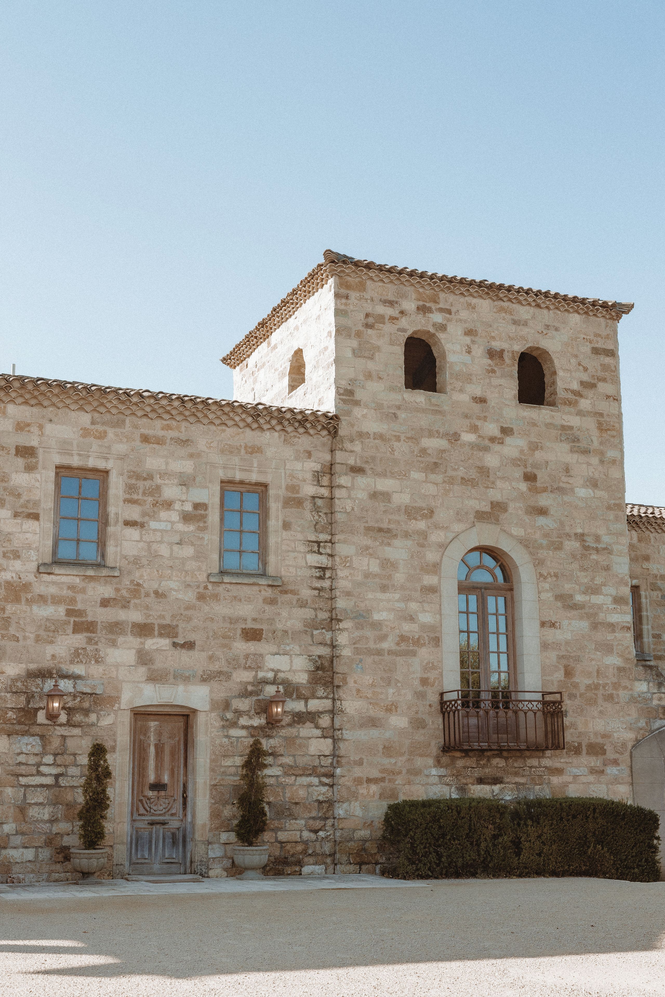 A stone building with a traditional design, featuring a central entrance with a door and windows, and a small balcony with a wrought iron railing.