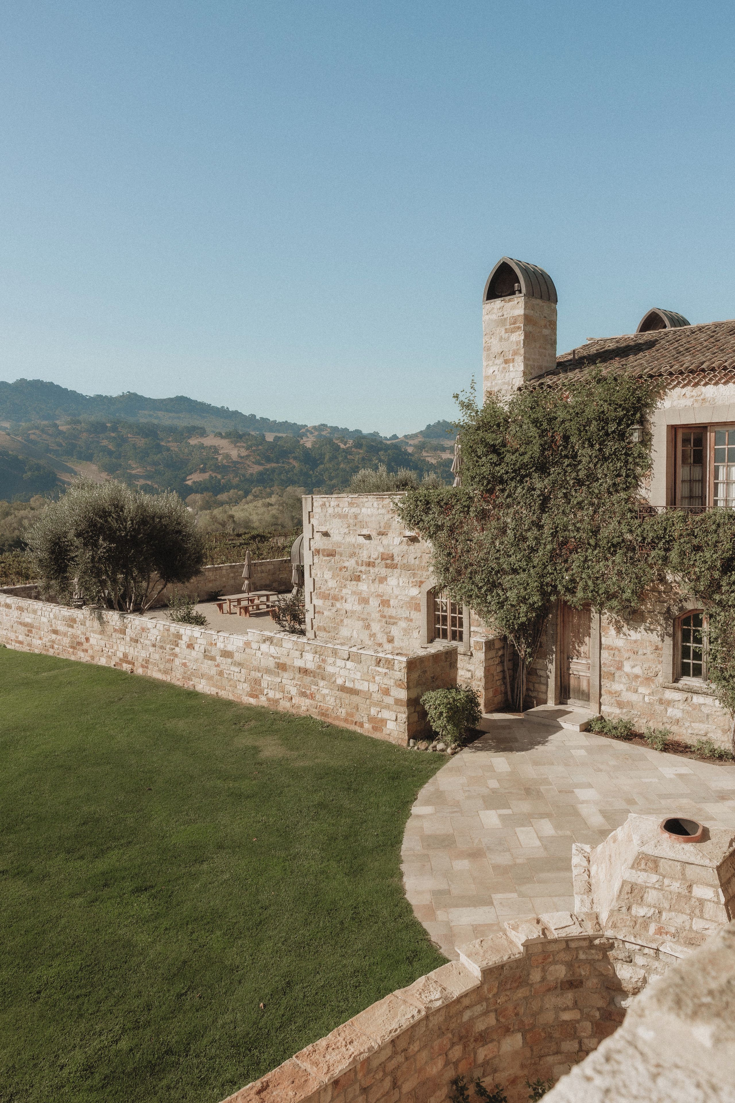 A stone building with a tower, a paved patio, and a view of hills.