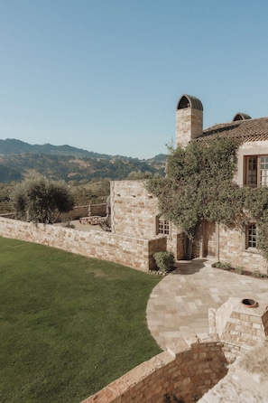 Un edificio de piedra con una torre, un patio empedrado y vistas a colinas.