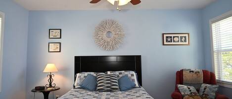 A bedroom with a bed, a ceiling fan, a lamp, a chair, and wall decorations.