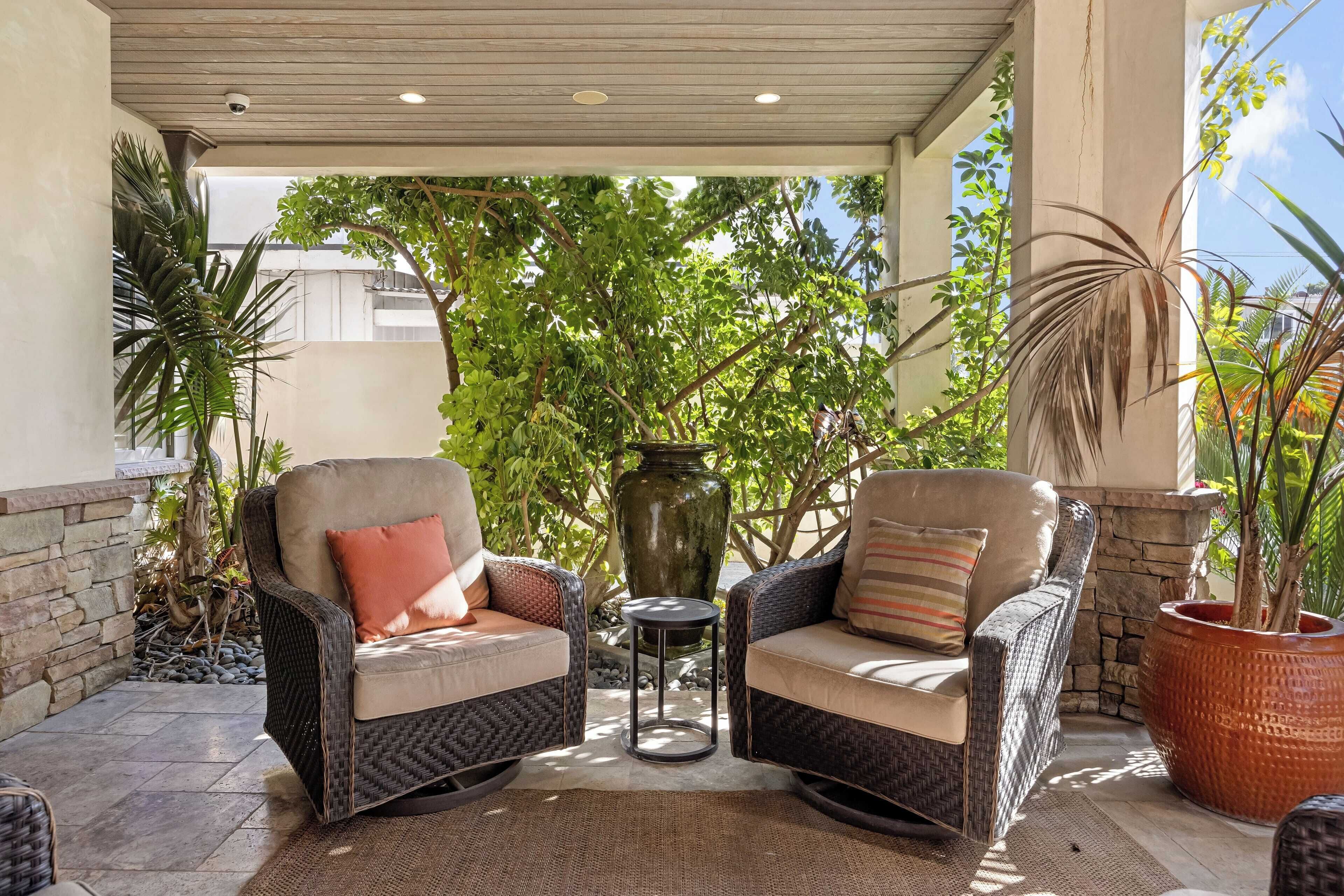 A covered patio area with wicker furniture, a large vase, and lush greenery.