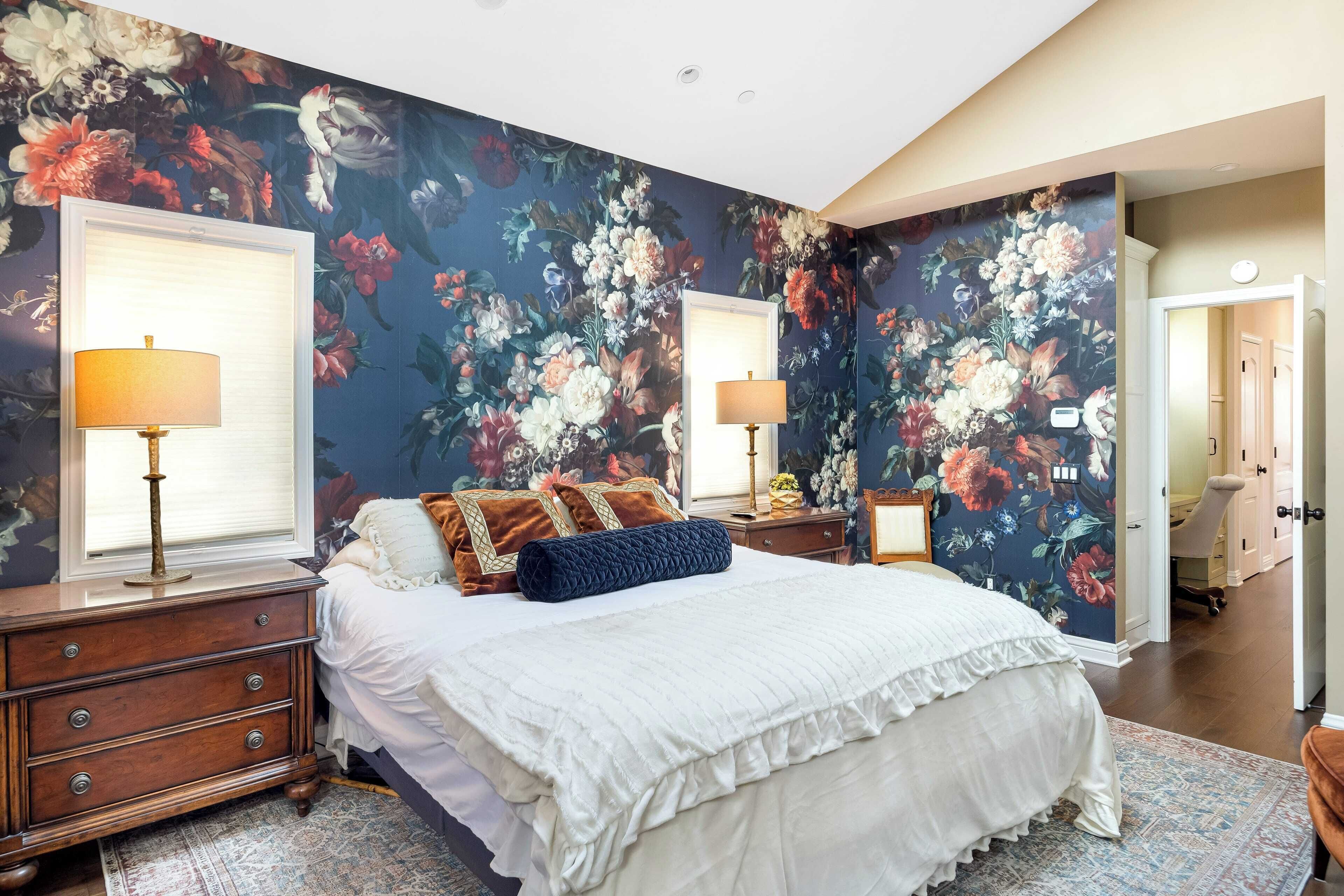 Ein Schlafzimmer mit floraler Tapete, einem großen Bett mit weißer Bettwäsche, zwei Nachttischen mit Lampen und einer Holzkommode.