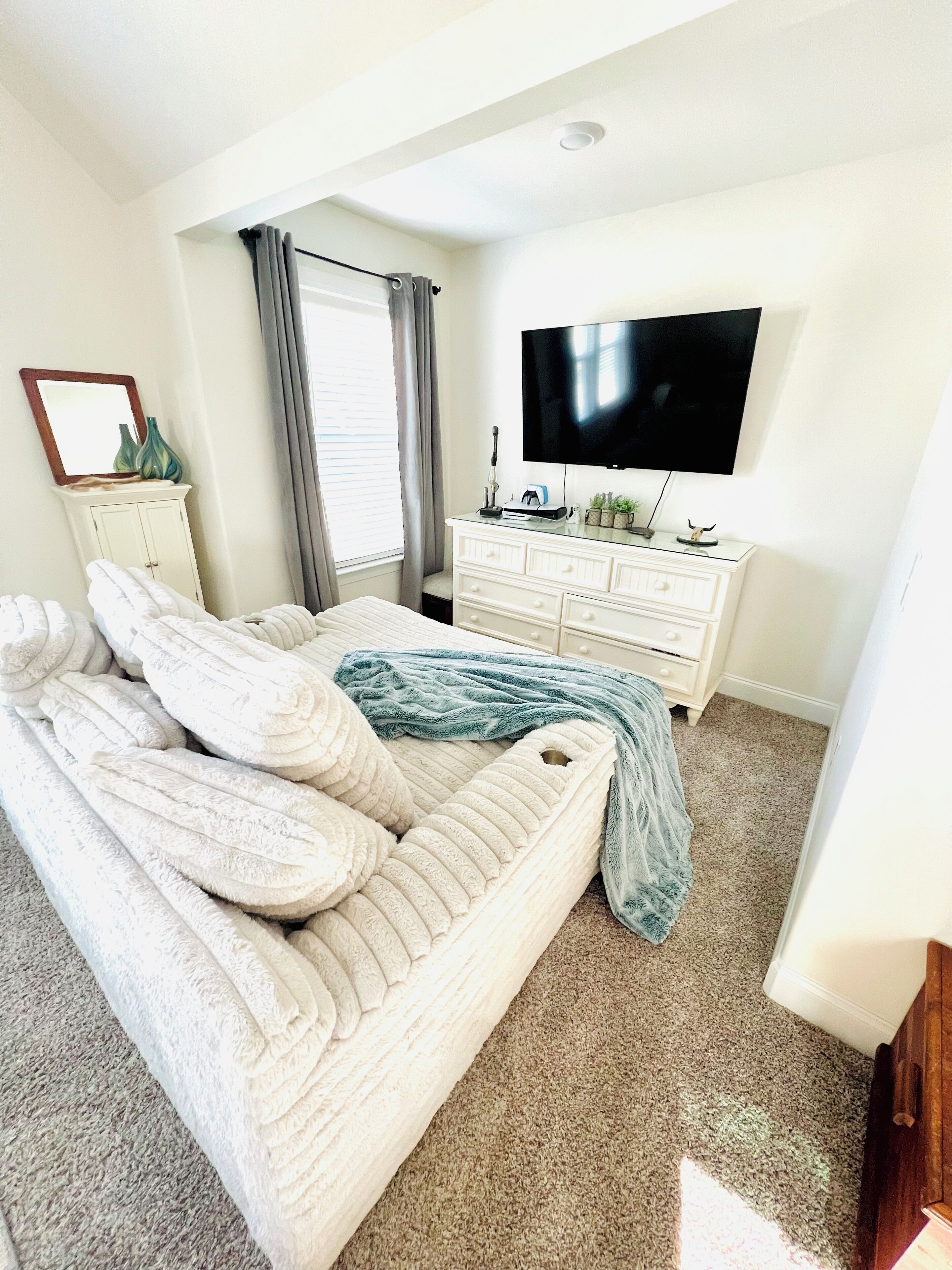 A bedroom with a large bed, a dresser, a flat-screen TV, and a window with curtains.
