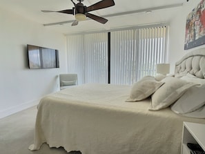 Primary bedroom with upholstered tufted headboard, crisp bedding, dresser, and large windows with vertical blinds - Elegant 2-Bedroom Beachfront Condo in Sandy Cove (Sarasota)