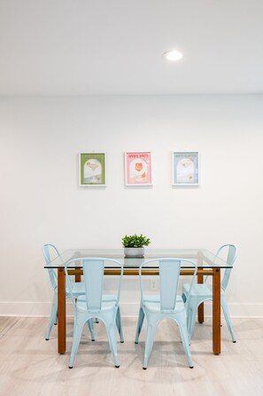 Un comedor con una mesa de vidrio y cuatro sillas azul claro, una planta en maceta sobre la mesa, y tres carteles enmarcados en la pared.