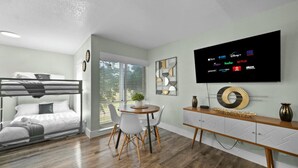 Una habitación moderna con literas, una zona de comedor con mesa y sillas, un televisor que muestra servicios de streaming y un elemento decorativo en la pared.