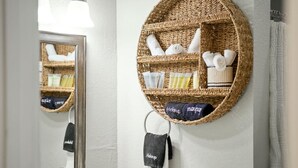 Un baño con un estante redondo de mimbre que contiene artículos de aseo y toallas, un espejo y un perchero con una toalla que dice 'Maquillaje'.