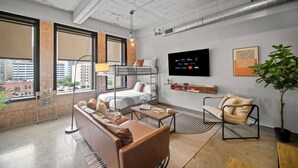 A modern hotel room with a large window, a bunk bed, a sofa, a coffee table, and a TV mounted on the wall.