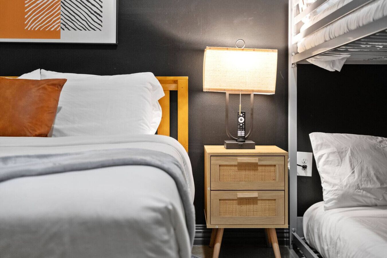 A hotel room with a bed, a bedside table with a lamp, a power outlet, and a bunk bed.