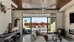 A modern living room with a large window, a ceiling fan, and a TV. - Windsong | Modern compound w pool & spa (Yucca Valley)