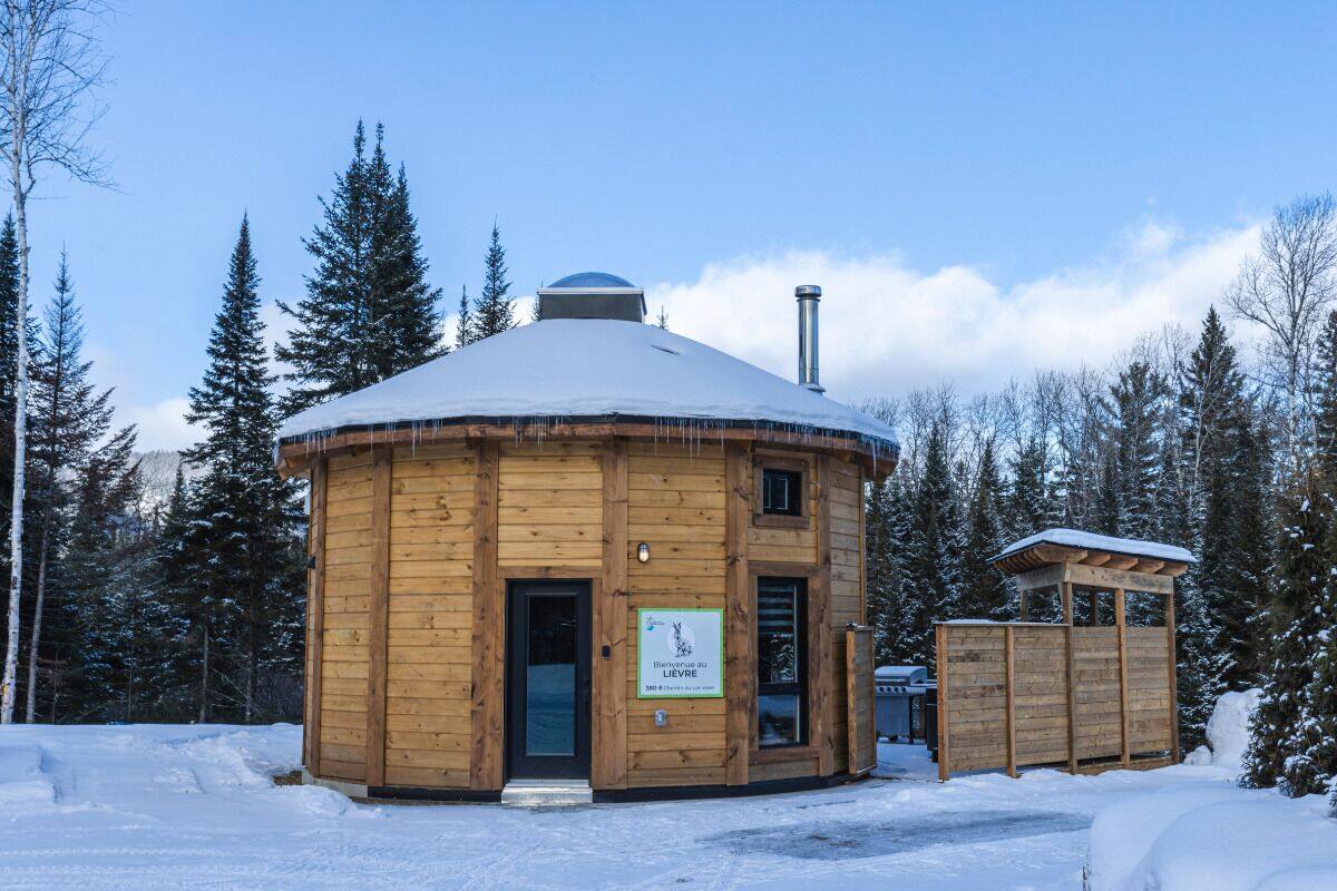 A wooden yurt with a snow-covered roof, surrounded by trees.