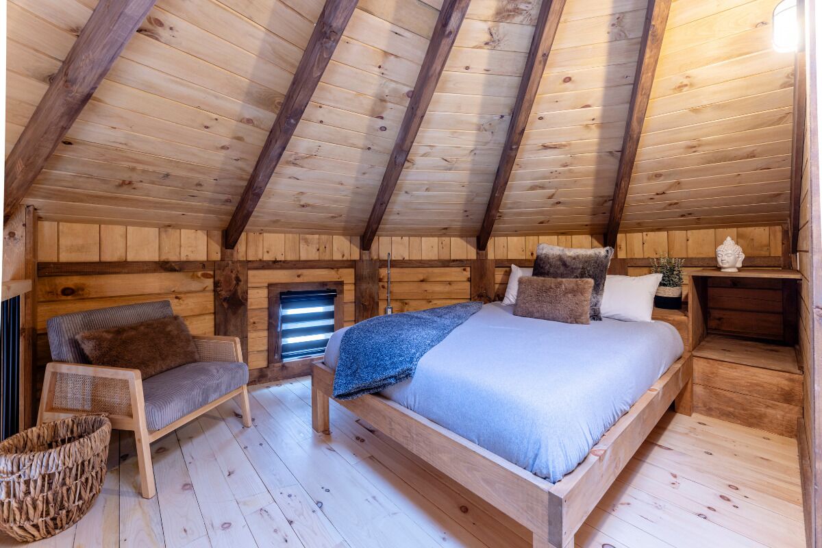 Ett mysigt sovrum på vinden med ett träpanelat tak, en säng med kuddar, en stol och ett litet bord med en dekorativ detalj.