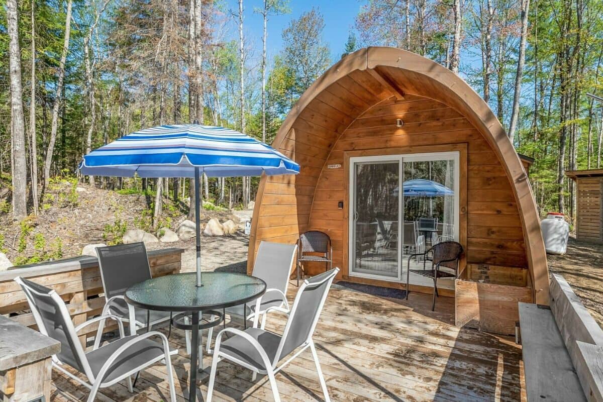 A wooden cabin with a glass door, a round table with chairs, and a blue-striped umbrella.