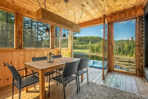 Una cabaña de madera con mesa y sillas, ventanas amplias y vistas a un lago y un bosque.