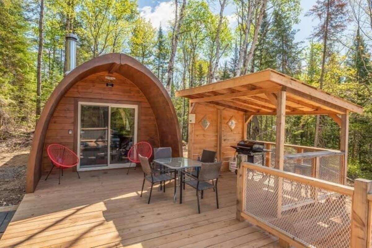 A wooden cabin with a covered patio, a dining area with chairs, and a grill.