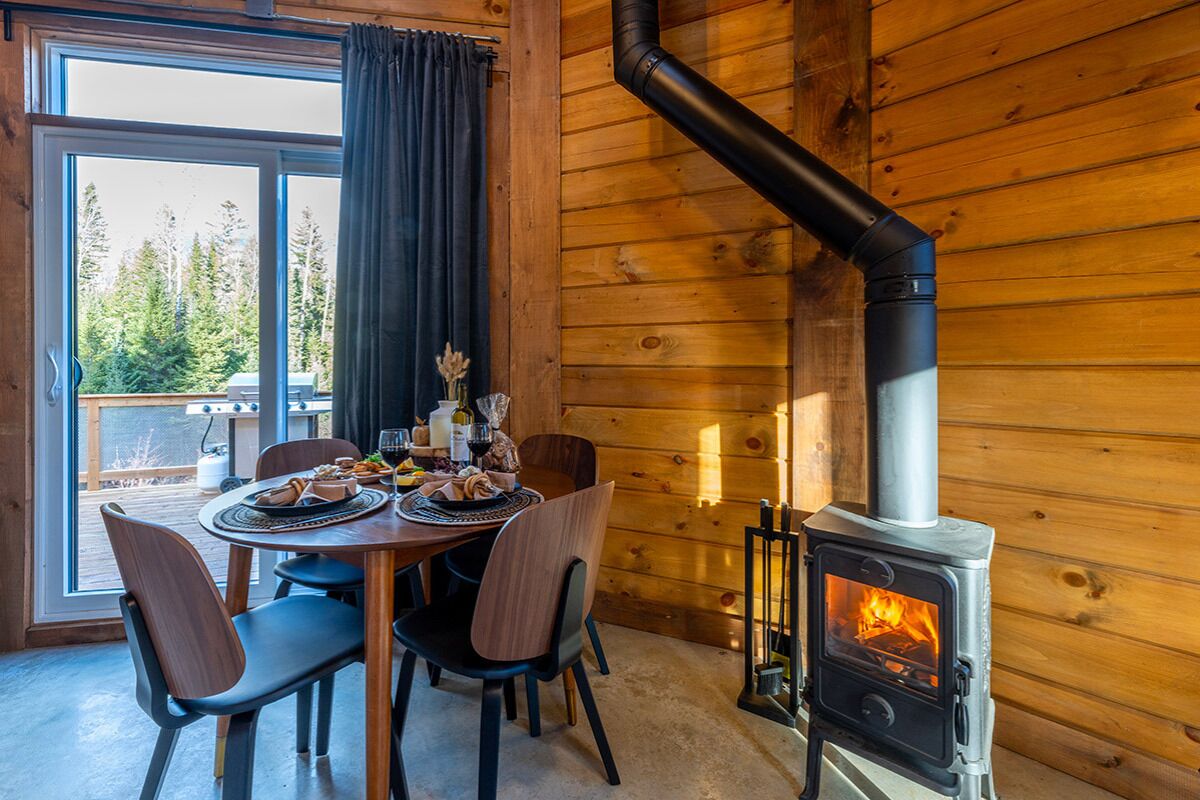 A wooden cabin interior with a dining table set for two, a wood-burning stove, and a view through a large window.