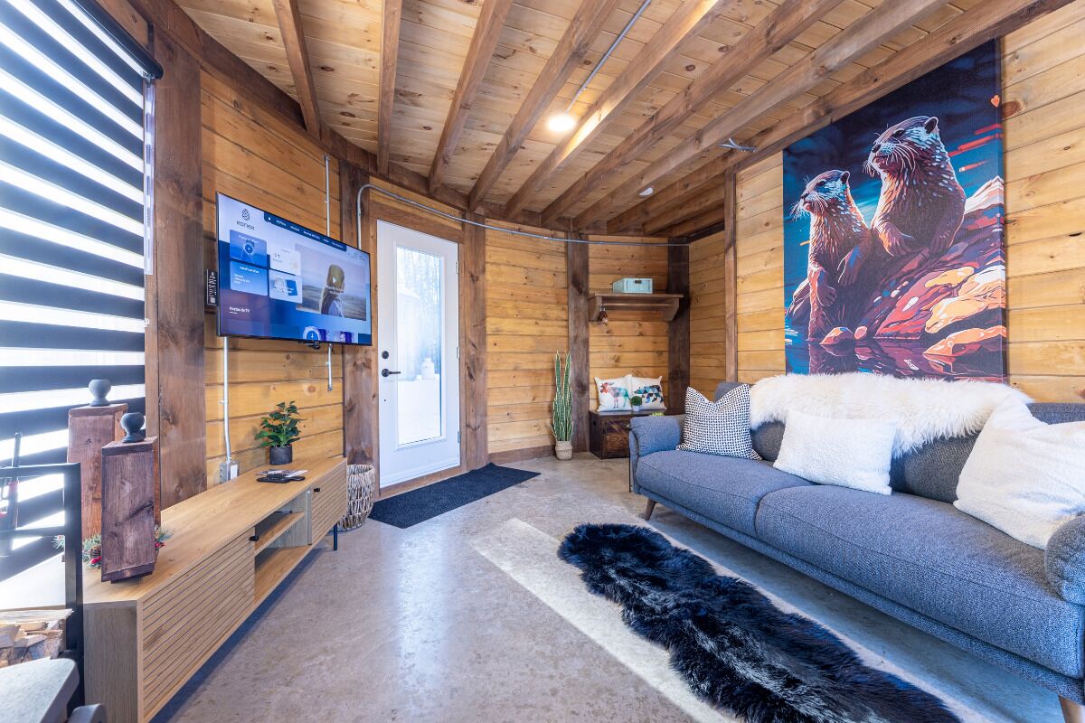 A cozy cabin interior with a wooden ceiling, a mounted TV, a sofa with a fur throw, and a painting of otters.