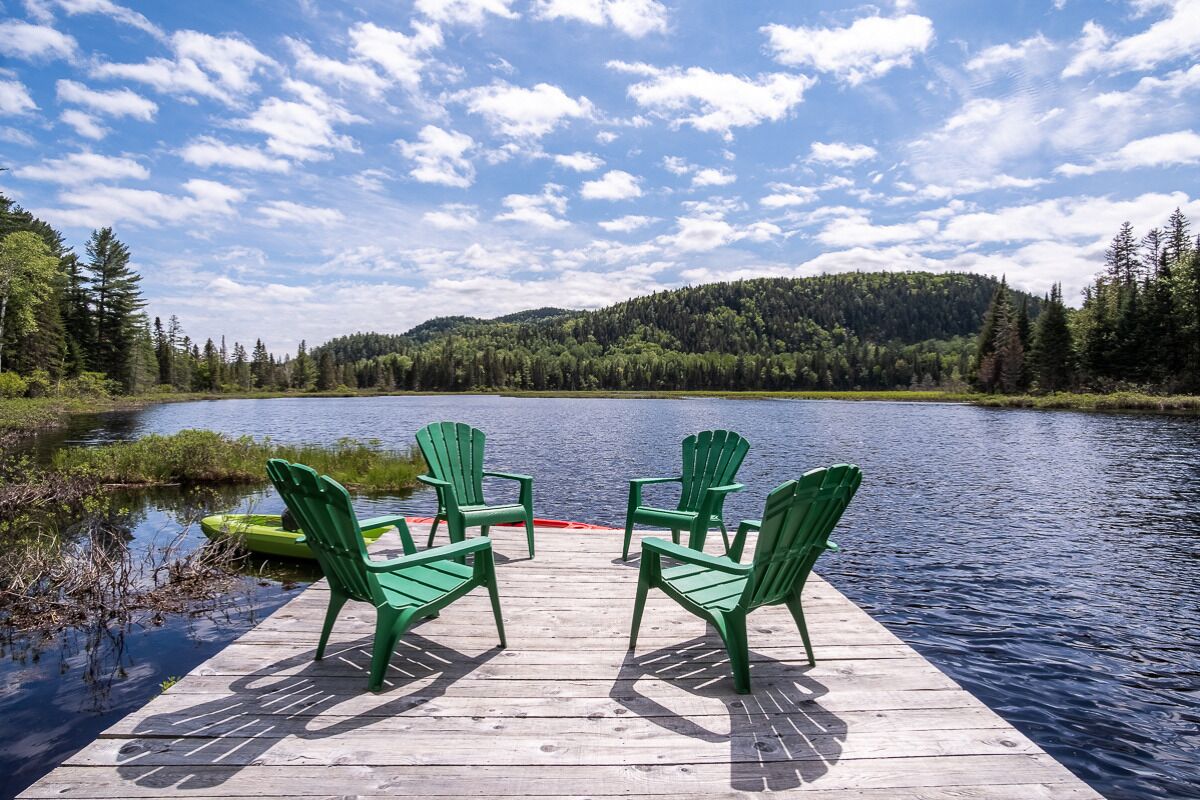 A wooden dock with four green Adirondack chairs facing a calm lake, surrounded by dense forest and hills under a partly cloudy sky.
