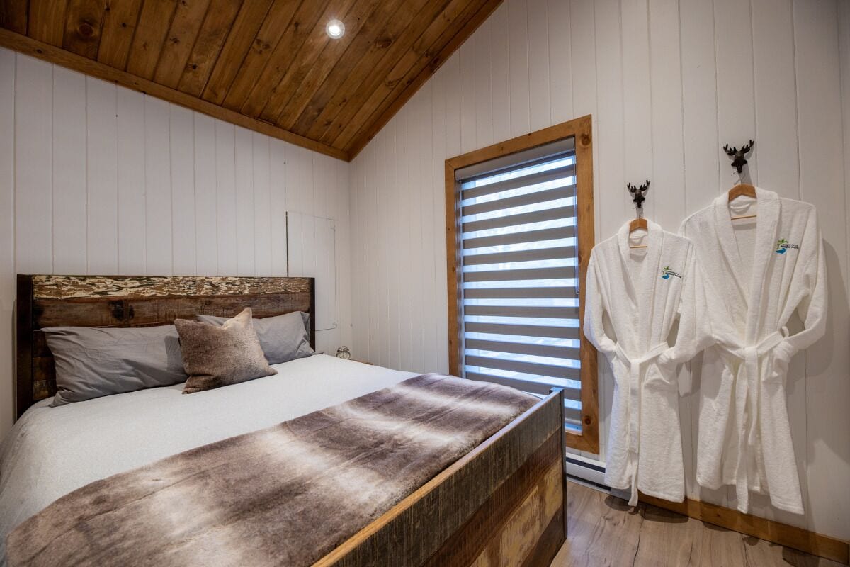 A bedroom with a wooden headboard, a large bed with a fur throw, two white robes on a rack, and a window with blinds.
