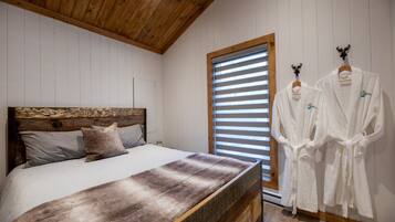 A bedroom with a wooden headboard, a large bed with a fur throw, two white robes on a rack, and a window with blinds.
