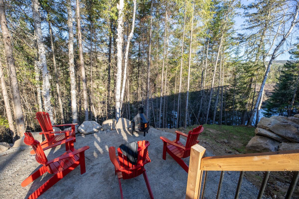 Une terrasse dotée de chaises Adirondack rouges, d’un poêle à bois et offrant une vue sur une zone boisée.