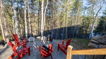 Une terrasse dotée de chaises Adirondack rouges, d’un poêle à bois et offrant une vue sur une zone boisée.