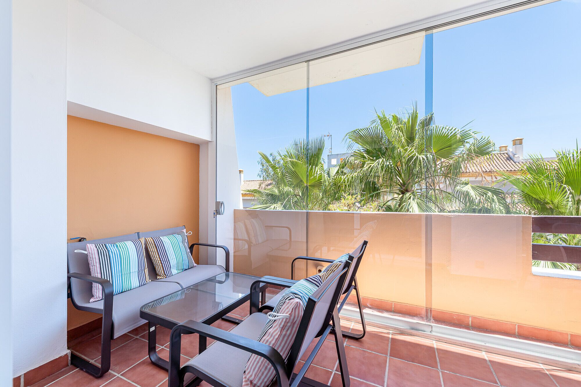 Ein Balkon mit einer Couch und zwei Stühlen, einem Glastisch und Blick auf Palmen und Gebäude.