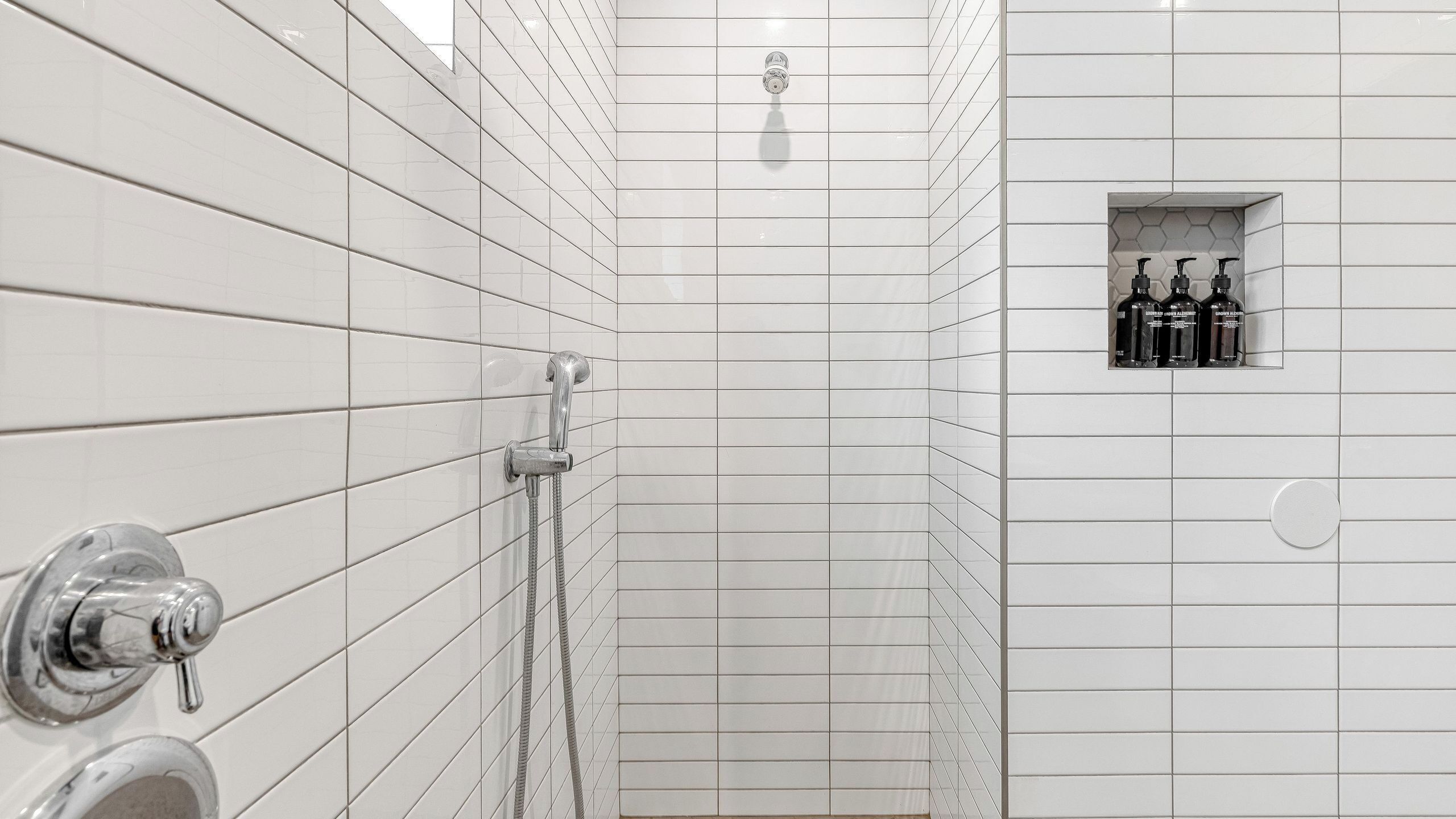 Une salle de bain avec des murs carrelés de carreaux blancs, un espace douche et une petite étagère contenant des articles de toilette.